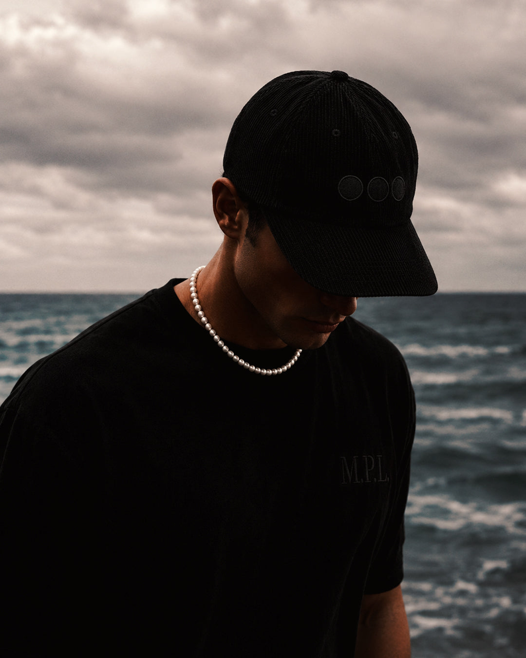 man walking beach in pearl necklace and black hat covering face